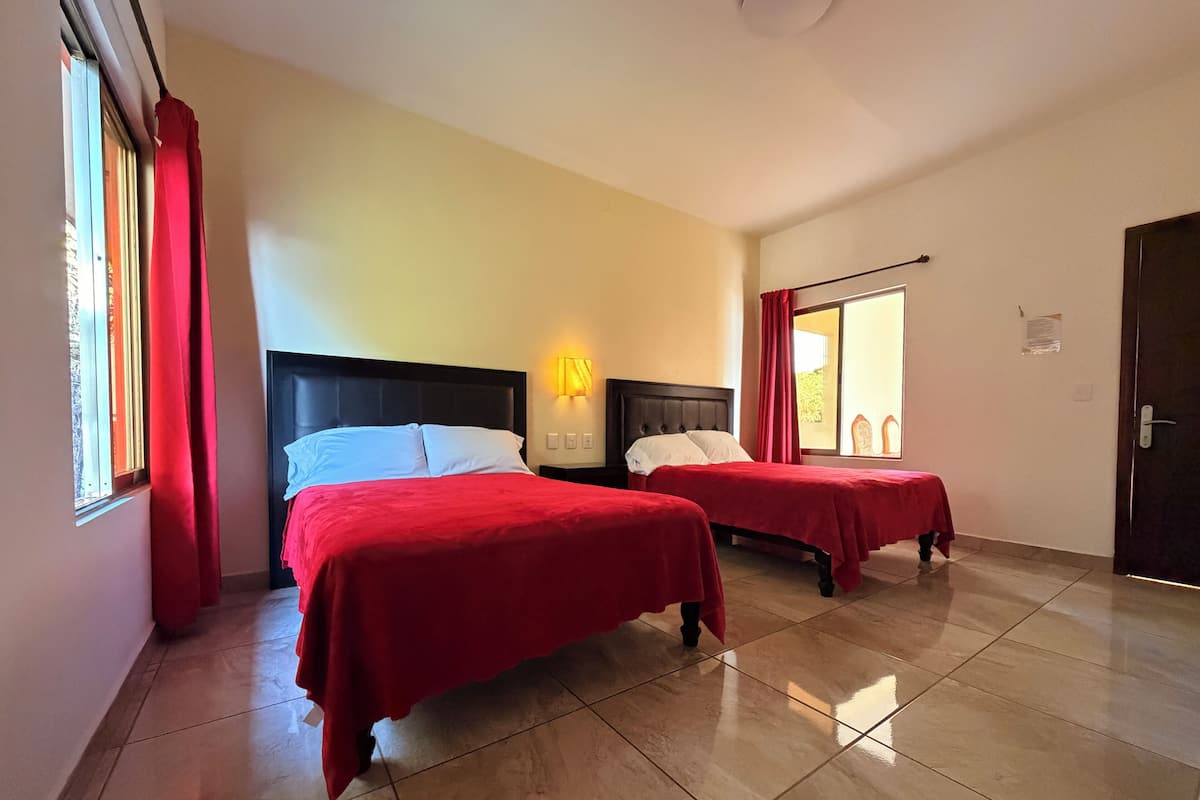 A hotel room with two beds, a window with red curtains, a wall-mounted light, and a door.