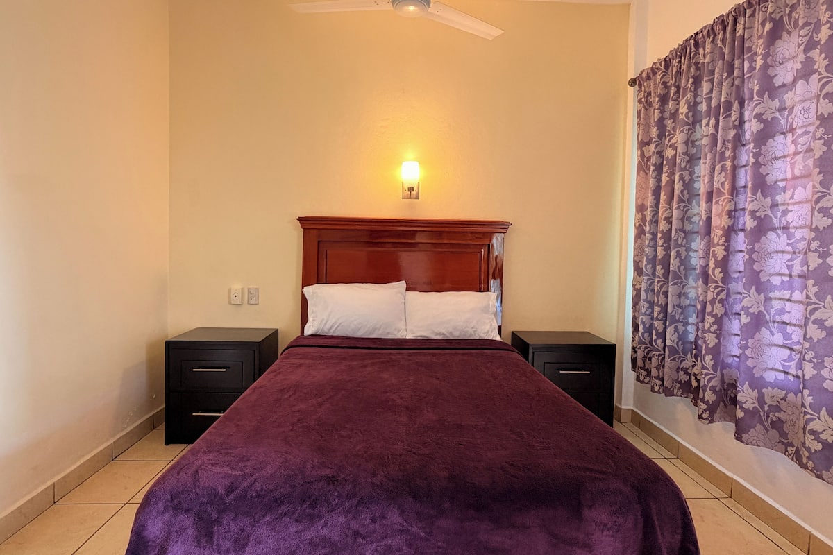A bedroom with a bed, nightstands, a ceiling fan, a wall-mounted light, and a floral curtain.