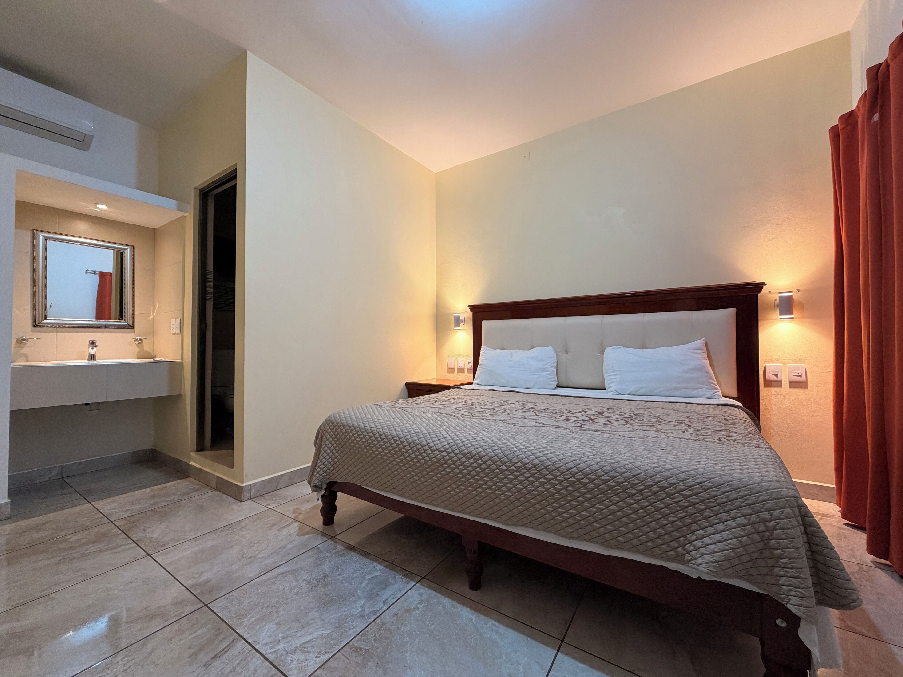Habitación de hotel con cama, baño con espejo y puerta que da a otra habitación.