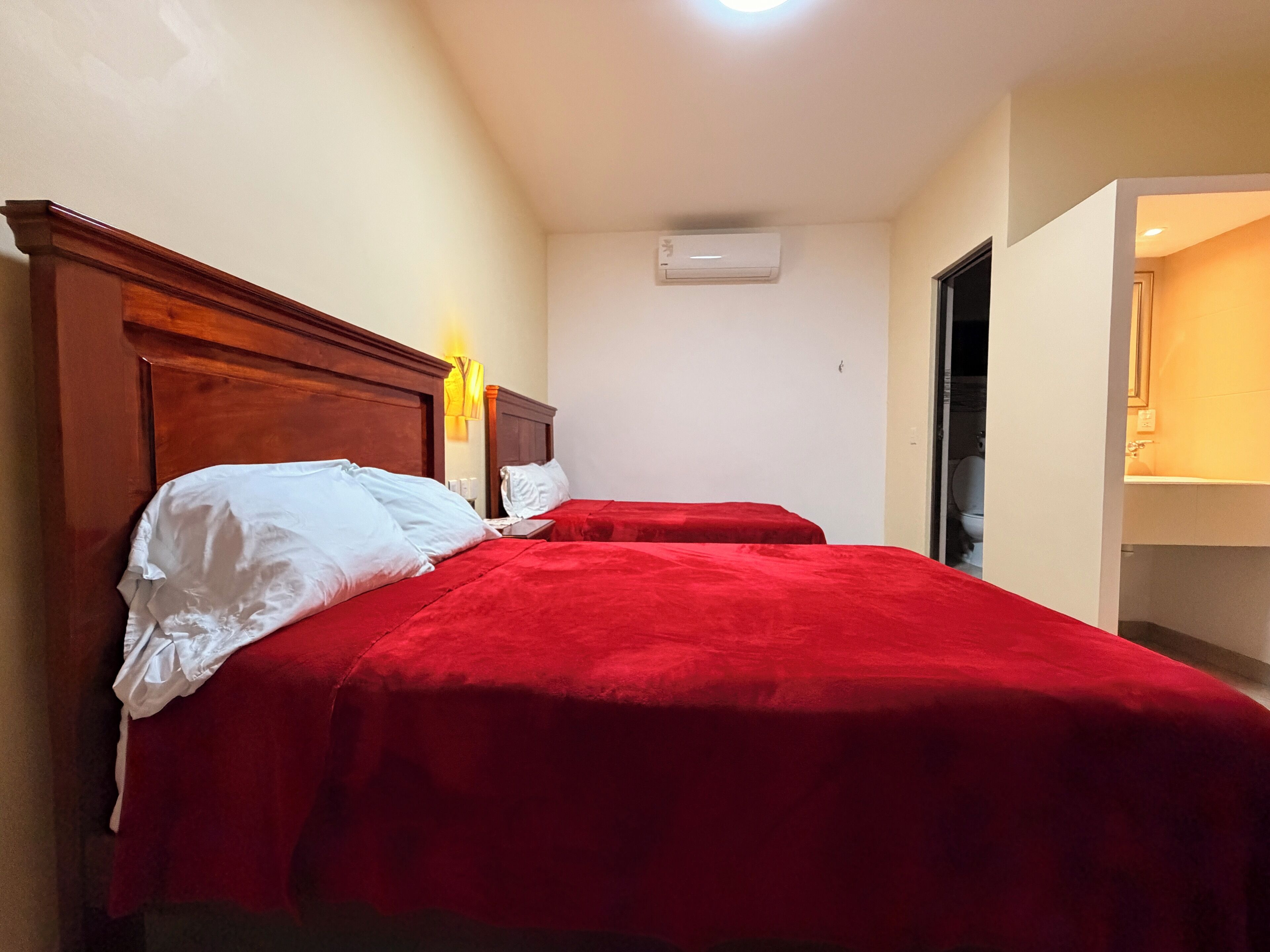 A hotel room with two beds, a wooden headboard, a wall-mounted air conditioner, and a bathroom visible through an open door.