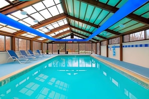 An indoor swimming pool with clear glass ceiling, blue lounge chairs, and a view of the outside.