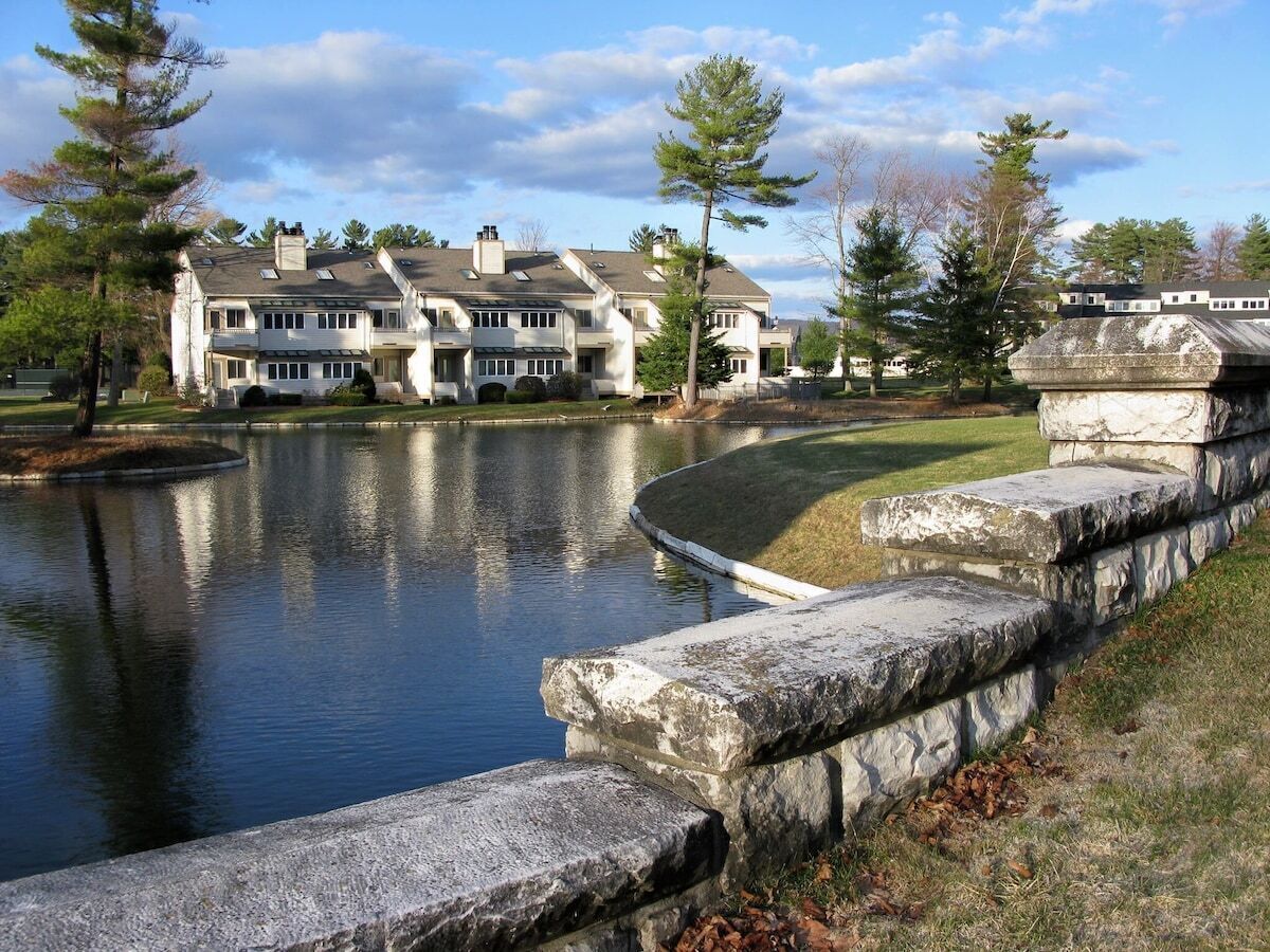 A large, multi-story house with a pond in front, stone steps, and a clear sky.