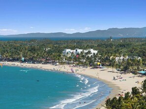 Una playa con aguas azul cristalino, una orilla arenosa y un telón de fondo de palmeras y montañas.