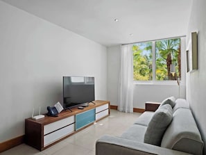 A modern living room with a flat-screen TV, wooden sideboard, grey sofa, and a large window with a view of greenery. - Oceanfront Apartment w/ Beachfront Pool in (Cabarete)