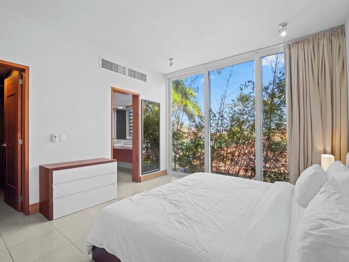 Une chambre dotée d’un grand lit, d’une coiffeuse avec un miroir, d’un banc en bois et d’une porte-fenêtre coulissante donnant sur un balcon avec vue sur les arbres.