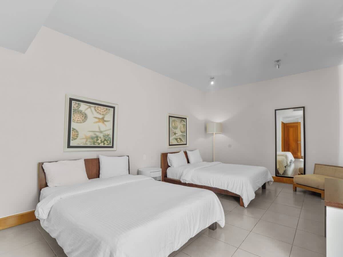A hotel room with two beds, a nightstand, a lamp, a chair, and two framed artworks on the wall.