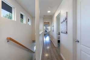 A hallway with a wooden handrail, a door with a picture, and a view into a room with a white couch. - Boise State Retreat | Walk to BSU & Downtown (Boise)