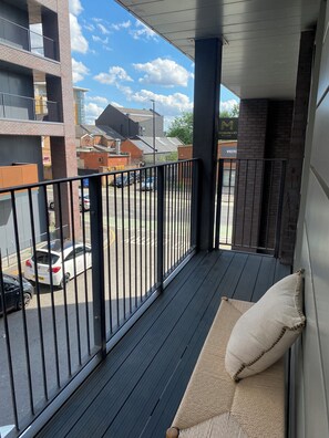 Ein Balkon mit einem Korbusofa und einem Kissen, einem Metallgeländer und Blick auf die Straße und die Häuser.