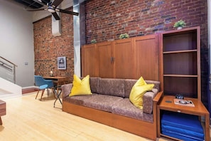 Ein modernes Wohnzimmer mit Backsteinwand, einem Holzsofa mit gelben Kissen, einem blauen Sessel und einem kleinen Tisch mit einer Uhr.