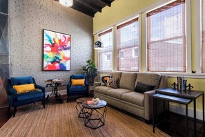 Ein Wohnzimmer mit Backsteinwand, einem farbenfrohen abstrakten Gemälde, zwei blauen Sesseln, einem beigen Sofa, einem kleinen runden Couchtisch und einem Beistelltisch mit Büchern.