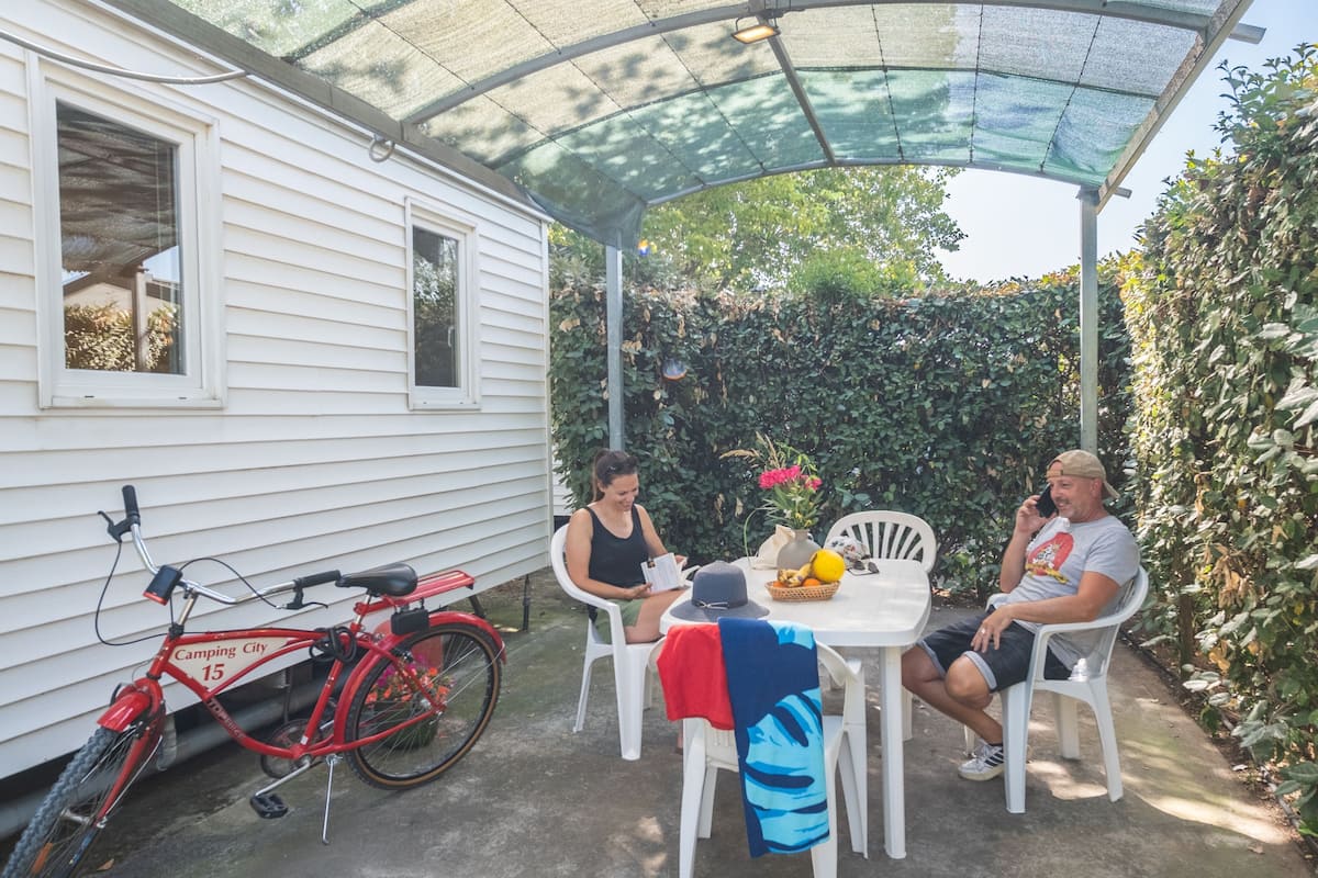 Um alpendre coberto com uma mesa e cadeiras, uma bicicleta vermelha e uma casa móvel branca.