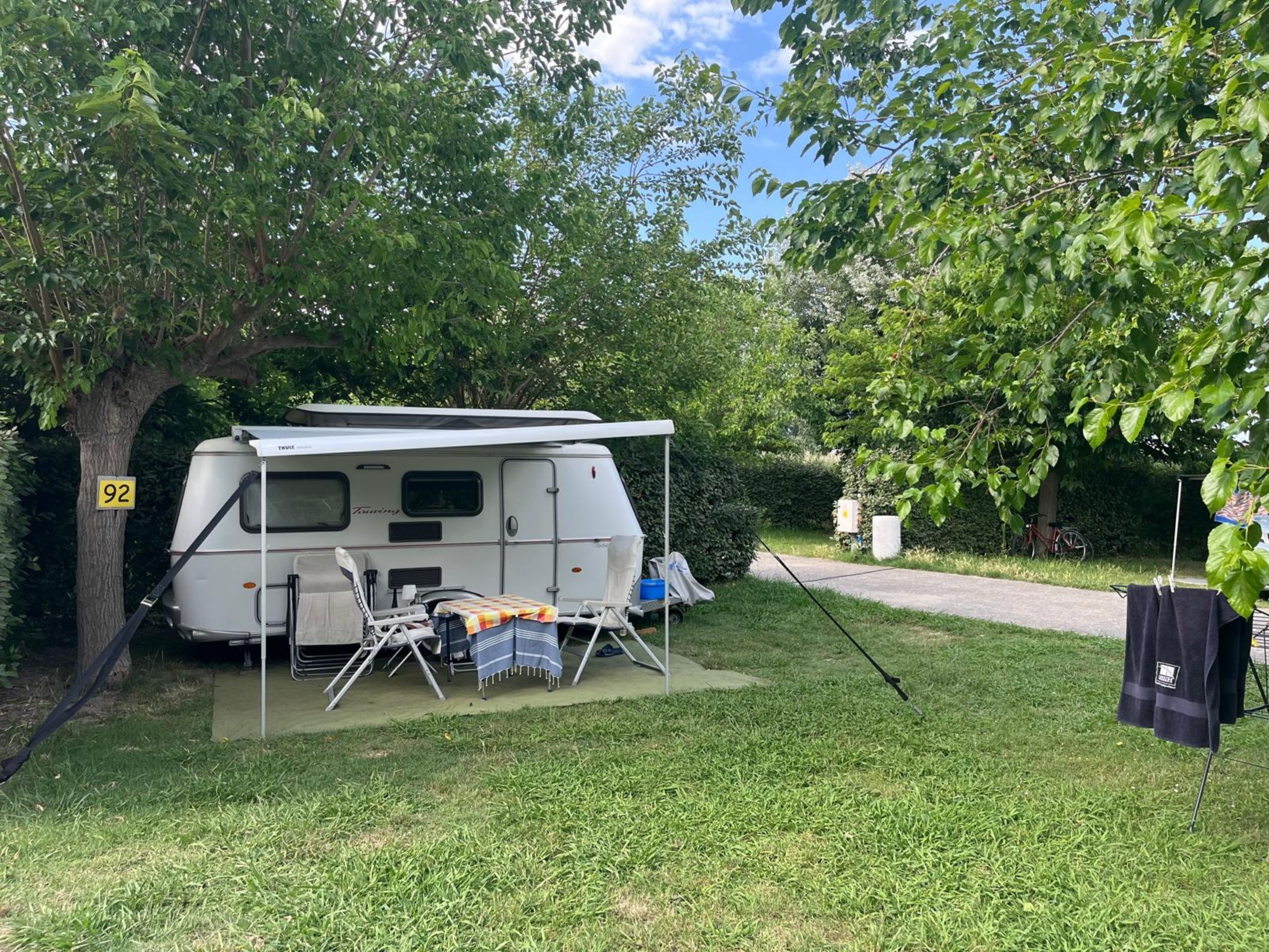 A white RV parked in a grassy area with a canopy, a table with a striped tablecloth, and a bicycle in the background.