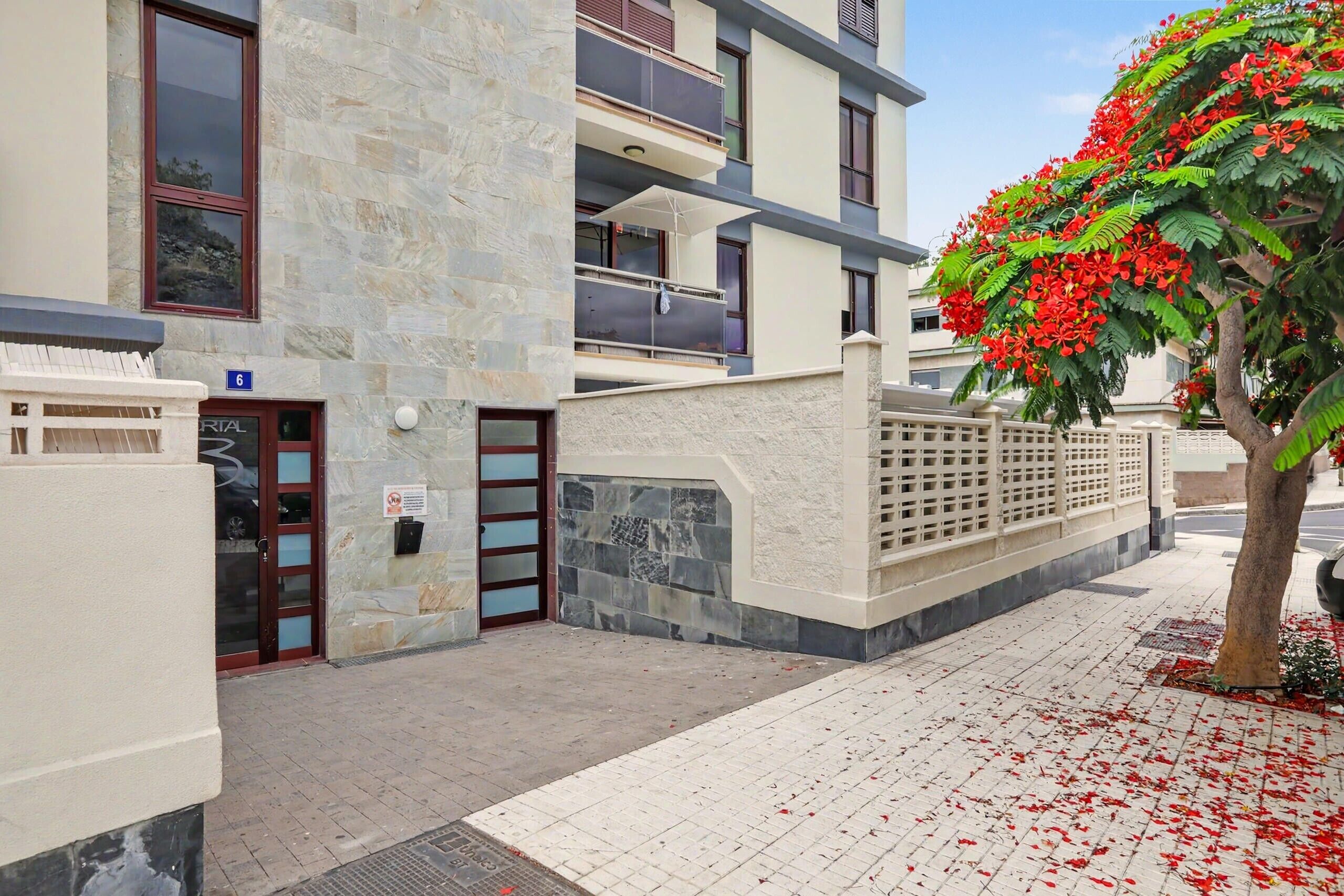 Un immeuble à plusieurs étages, doté d’une façade en pierre, d’une entrée sécurisée par un portail et d’un arbre aux fleurs rouges.