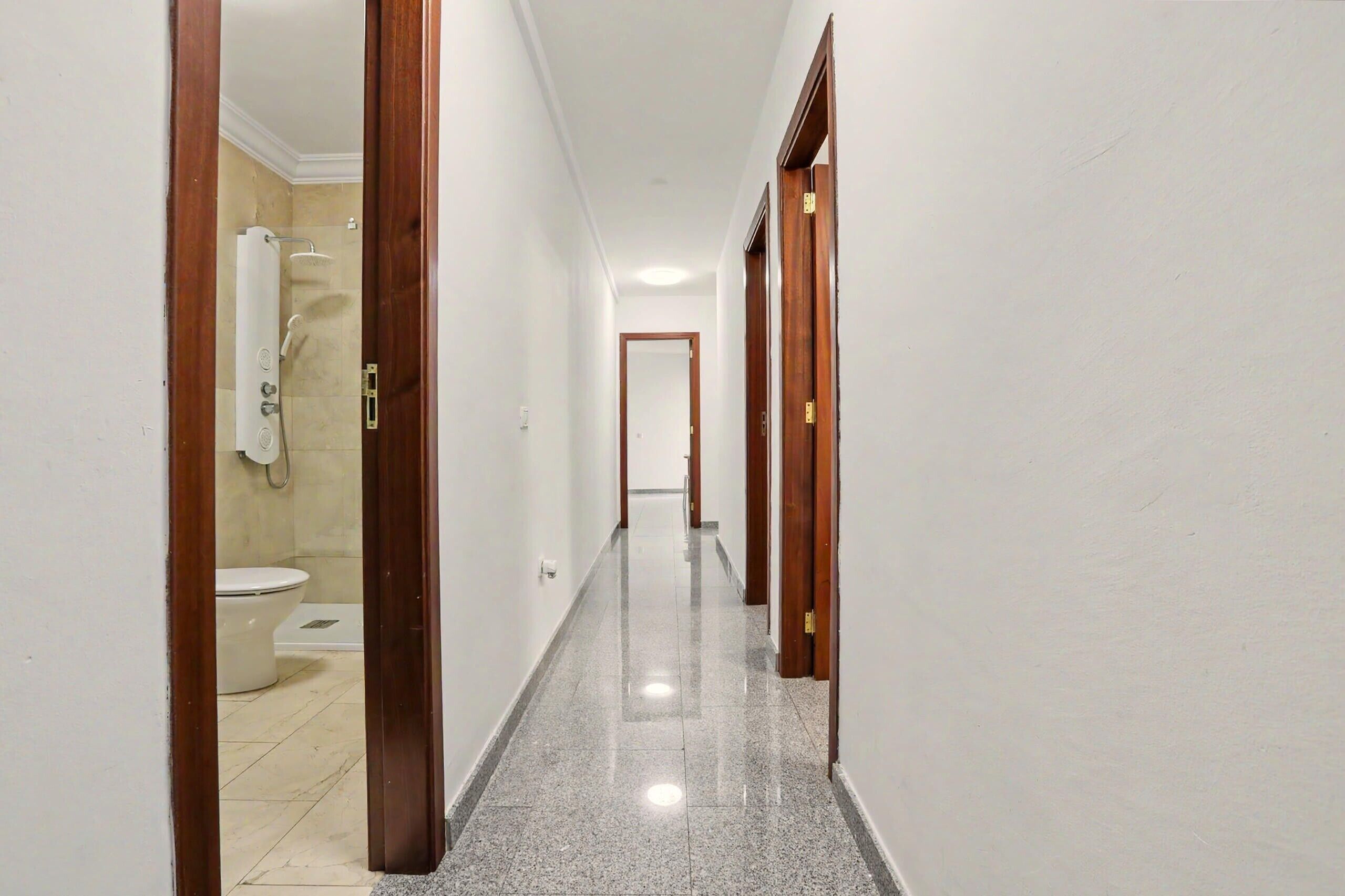 Un pasillo con un baño a la izquierda, puertas de madera a la derecha y piso de baldosas.
