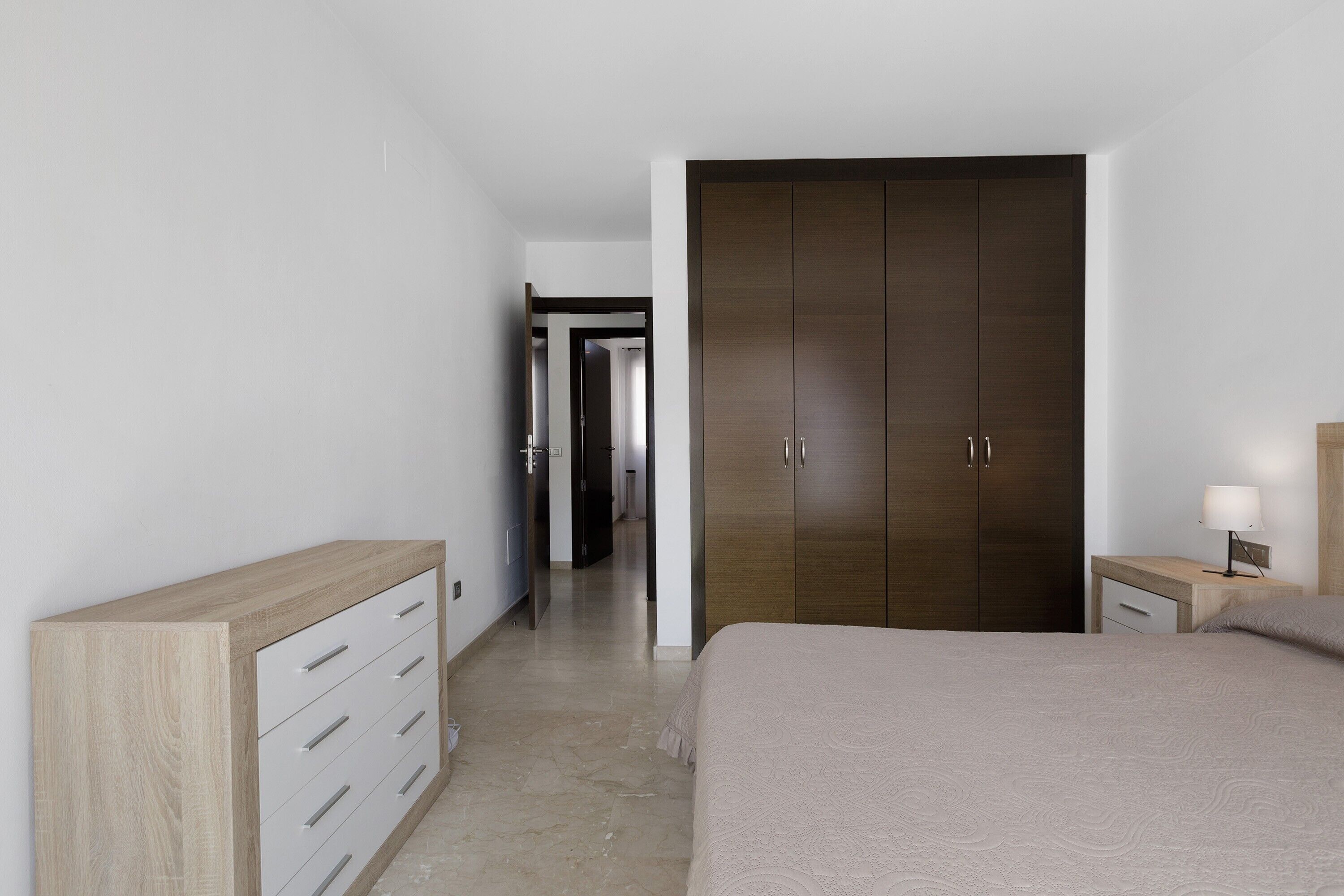 A bedroom with a bed, a wooden dresser, a wardrobe, and a bedside table with a lamp.
