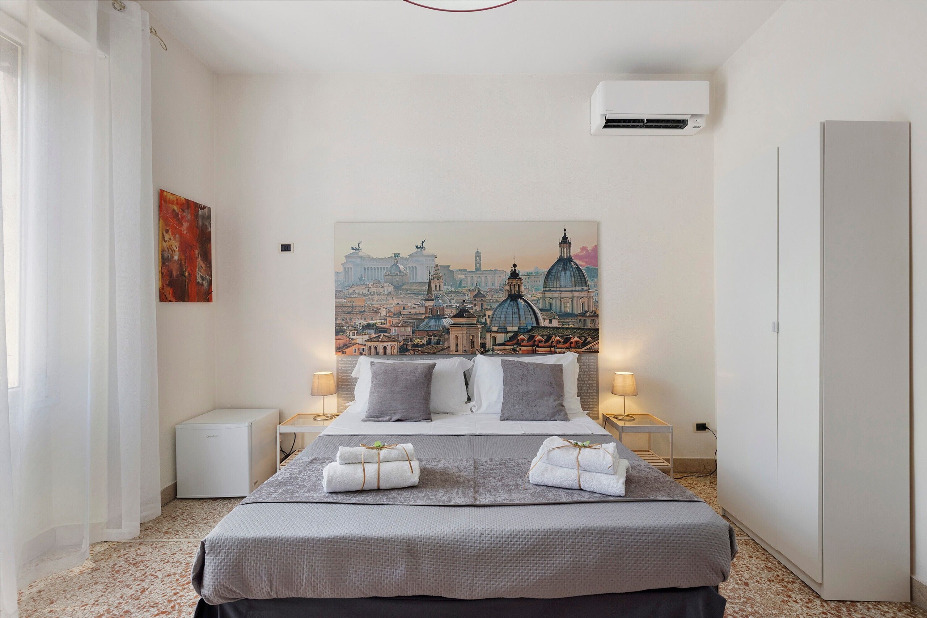 Une chambre d’hôtel avec un lit, un tableau représentant un paysage urbain, une table de chevet avec une lampe et un appareil de climatisation.