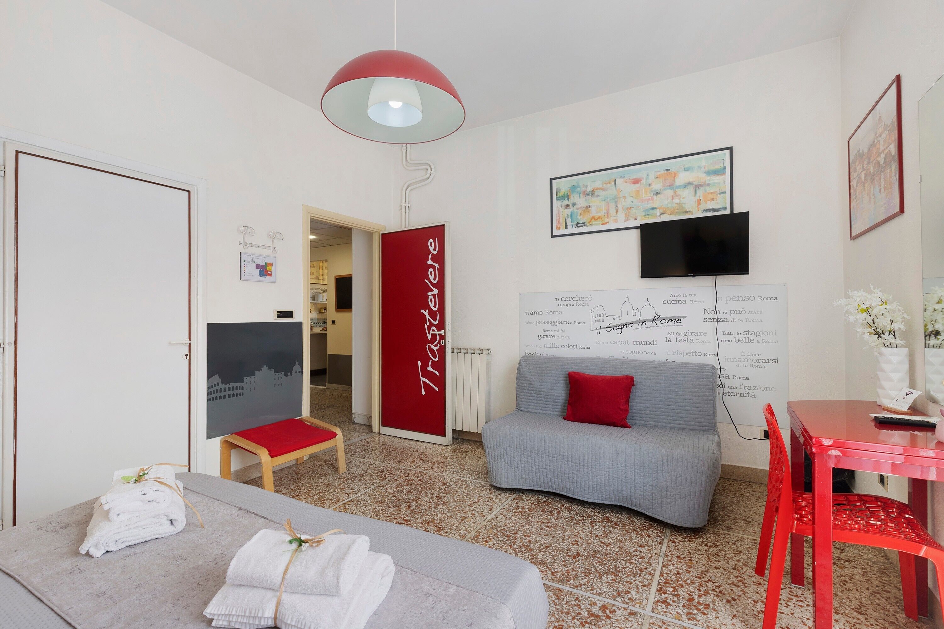 Habitación de hotel moderna con un sofá gris, un cojín rojo decorativo, una mesa roja y una silla roja. Se observa una pared blanca con una puerta roja y otra pared blanca con un televisor negro y un cuadro enmarcado.