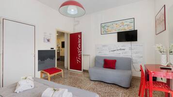 Habitación de hotel moderna con un sofá gris, un cojín rojo decorativo, una mesa roja y una silla roja. Se observa una pared blanca con una puerta roja y otra pared blanca con un televisor negro y un cuadro enmarcado.