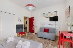 Habitación de hotel moderna con un sofá gris, un cojín rojo decorativo, una mesa roja y una silla roja. Se observa una pared blanca con una puerta roja y otra pared blanca con un televisor negro y un cuadro enmarcado.