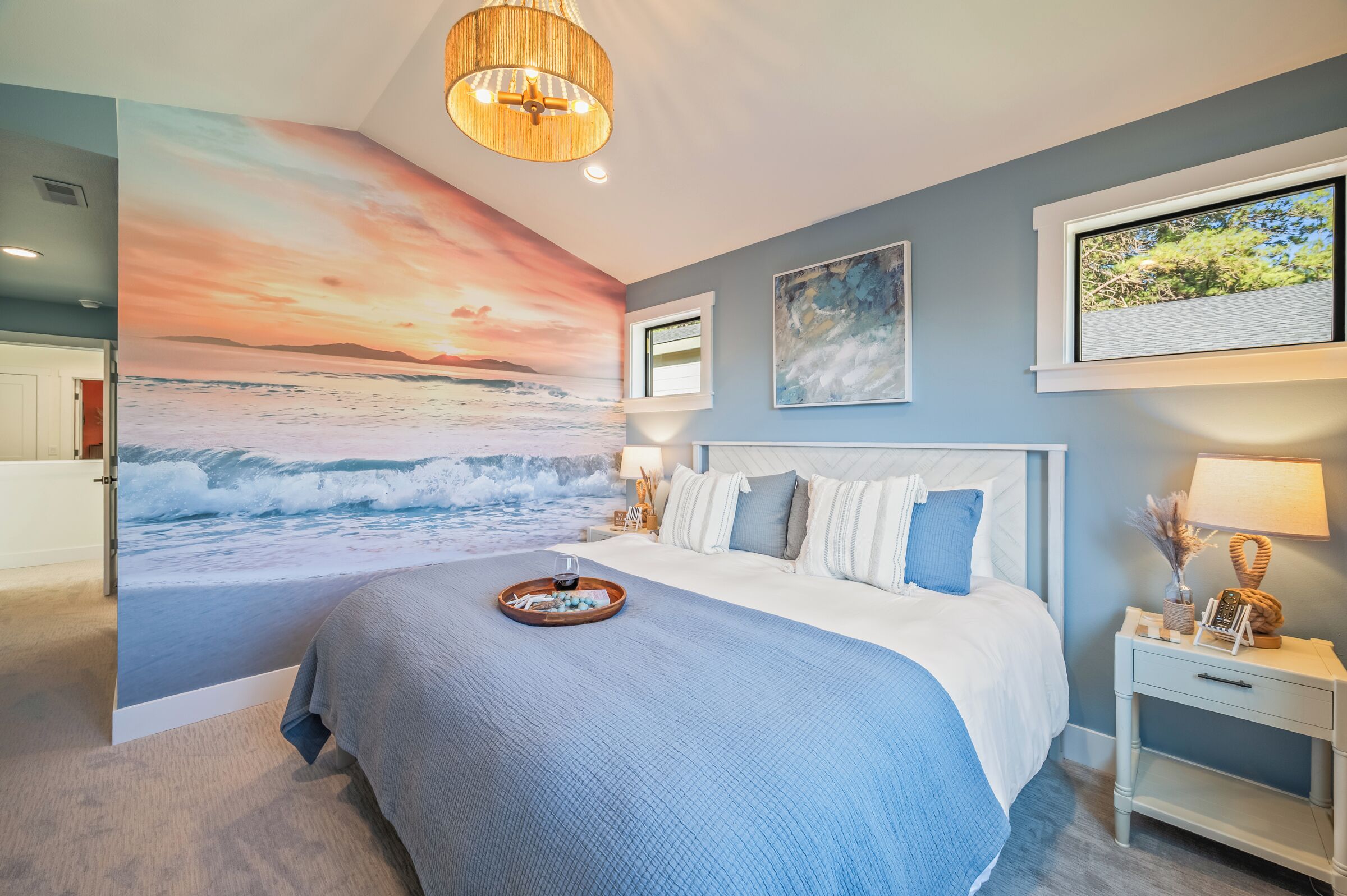 Ein Schlafzimmer mit einem großen Wandbild, das einen Sonnenuntergang über dem Meer zeigt, einem Bett mit blauer und weißer Bettdecke, einem Nachttisch mit Lampe und einem Fenster mit Blick auf Bäume.