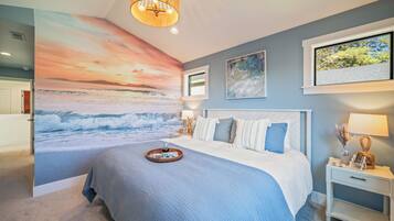 Une chambre avec une grande fresque murale représentant un coucher de soleil sur l’océan, un lit recouvert d’une courtepointe bleue et blanche, une table de chevet avec une lampe, et une fenêtre donnant sur des arbres.