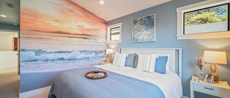 Quarto com um grande mural na parede retratando um pôr do sol sobre o oceano, uma cama com colcha azul e branca, uma mesa de cabeceira com abajur e uma janela com vista para árvores.