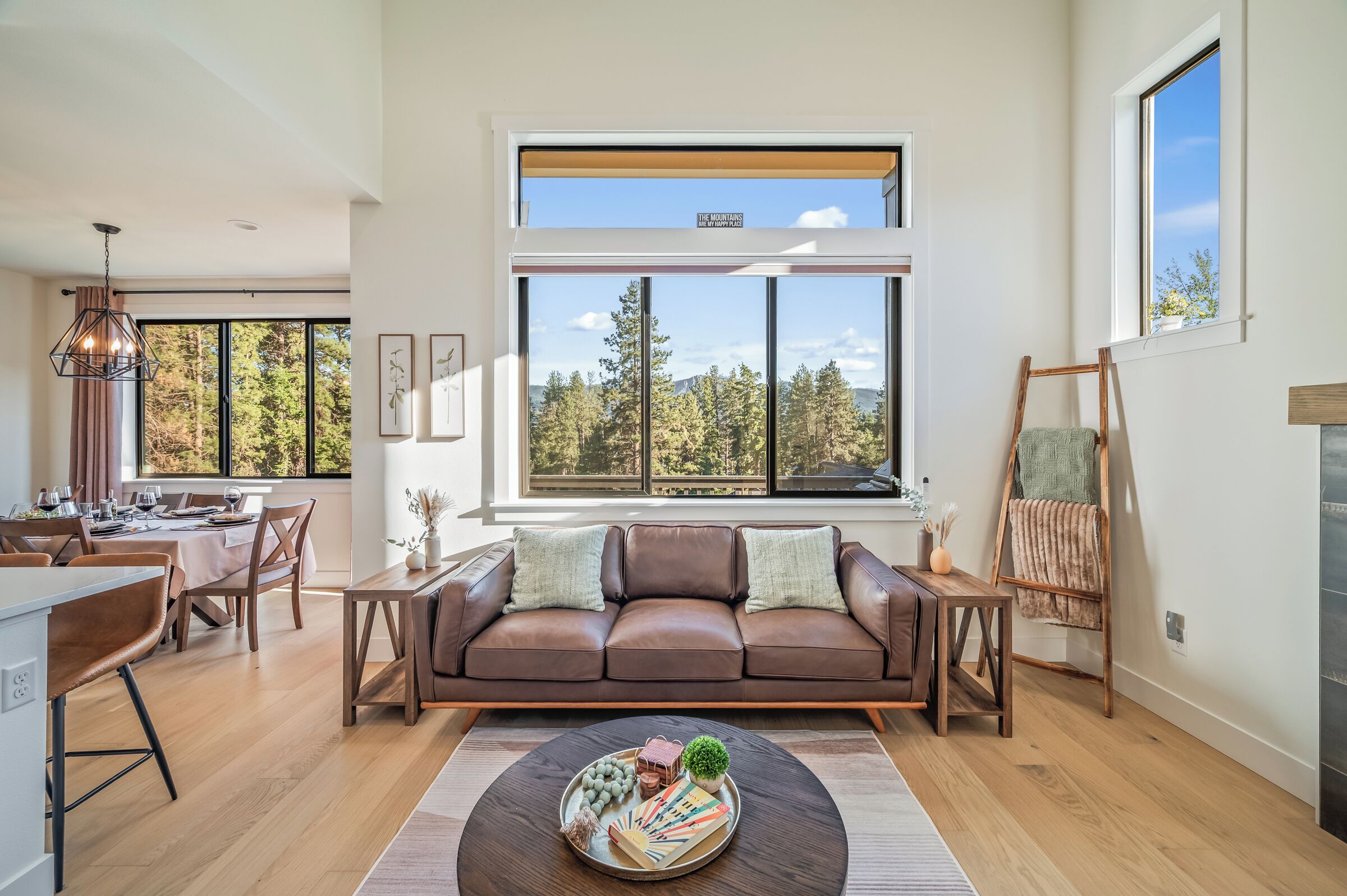 A modern living room with a brown leather sofa, a dining area with a table set for two, and large windows offering a view of trees and mountains.