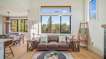 A modern living room with a brown leather sofa, a dining area with a table set for two, and large windows offering a view of trees and mountains.