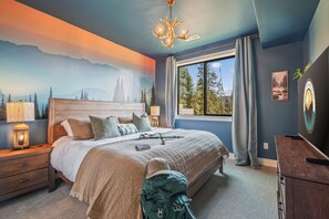 A bedroom with a large bed, a chandelier, a window with curtains, a nightstand with a lamp, and a wall mural. - Welcome to Outfitter Retreat, a mountain getaway within walking distance of Historic Downtown Cle Elum. Enjoy hiking, biking, ATV trails, golf, and year-round adventure. Each space has been designed to provide you with an unforgettable experience. (Cle Elum)