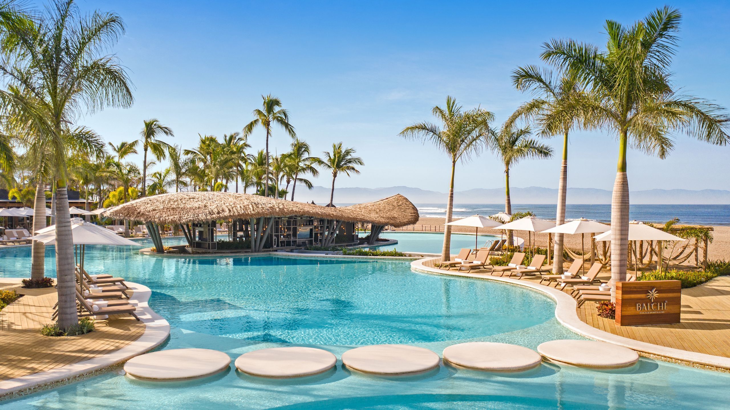 Uma área de piscina com espreguiçadeiras, palmeiras e uma estrutura com cobertura de palha.