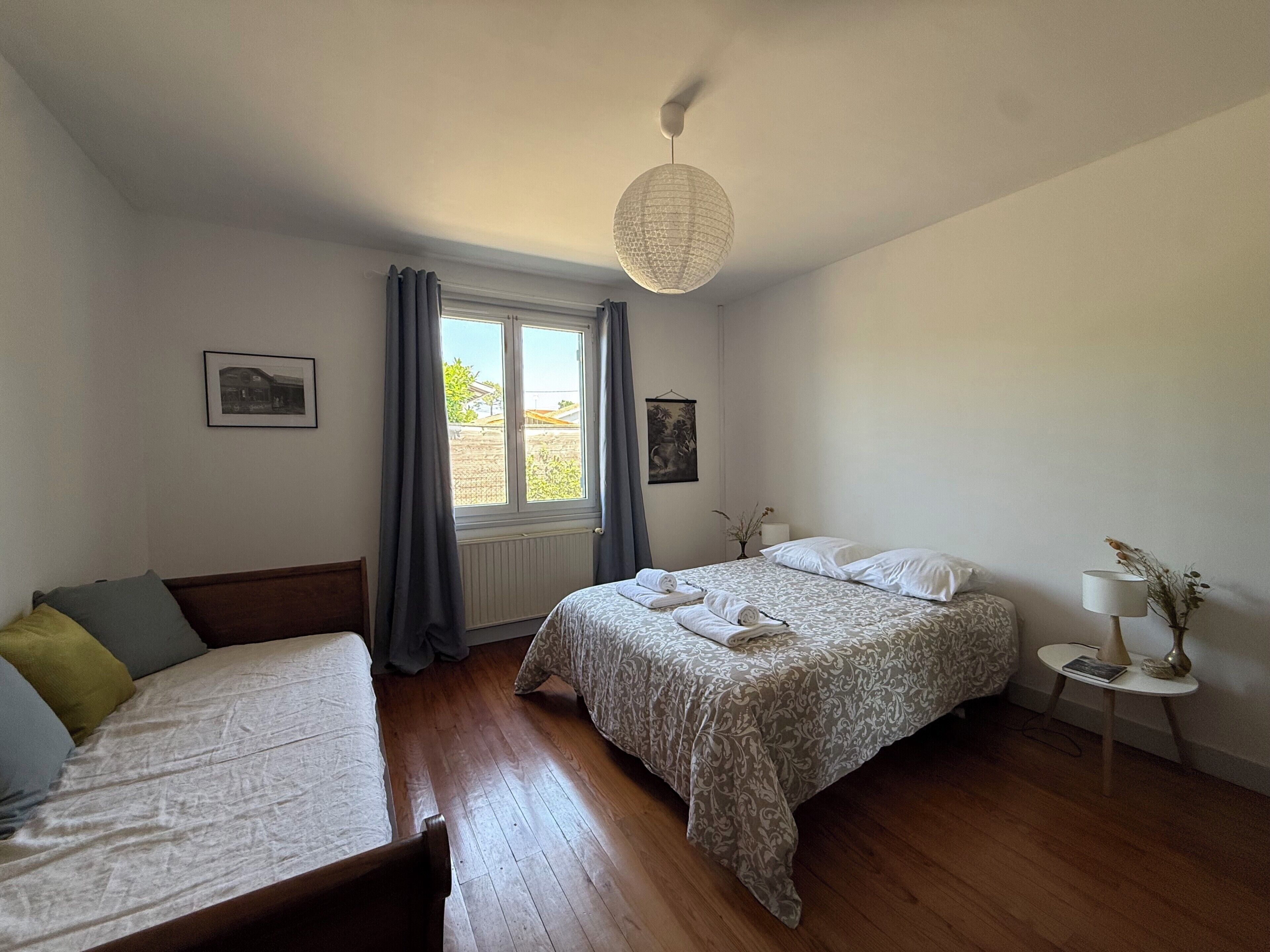 Een slaapkamer met twee bedden, een raam met gordijnen, een hanglamp en een klein nachtkastje met een lamp.