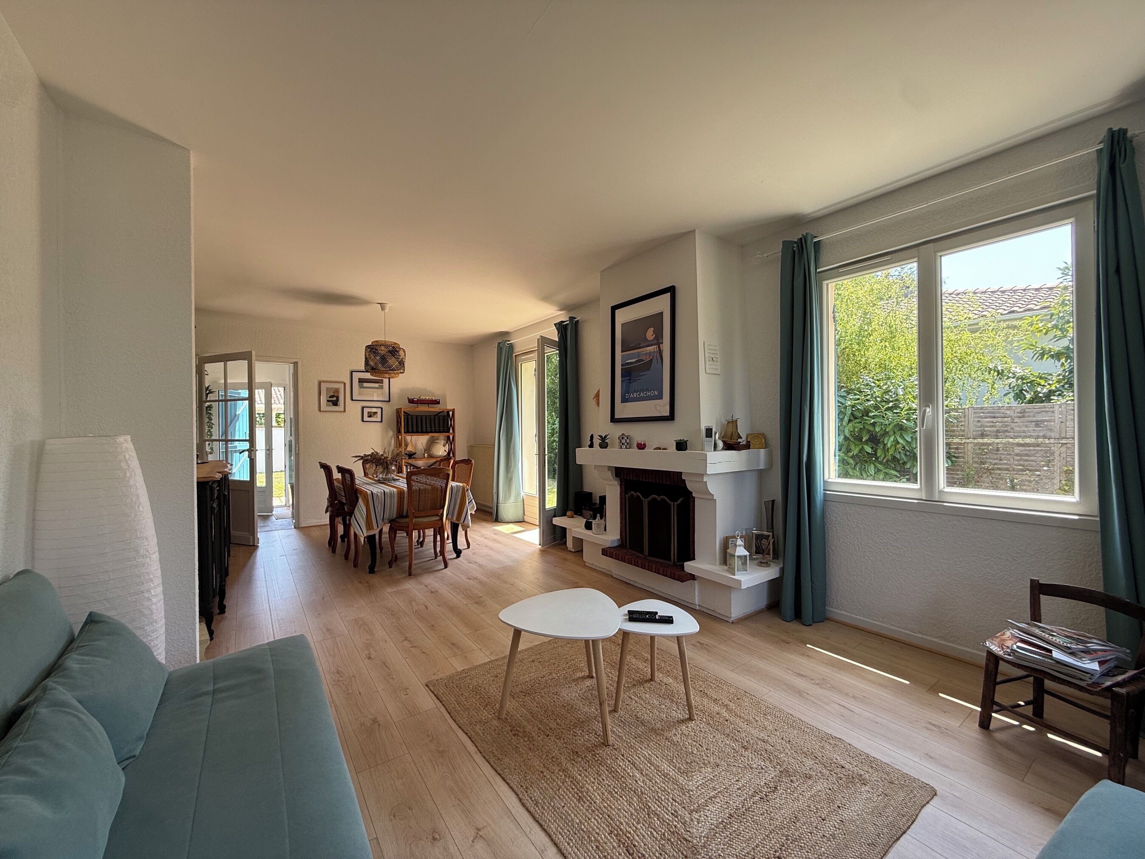 A living room with a sofa, a fireplace, a dining area with chairs and a table, and large windows with curtains.