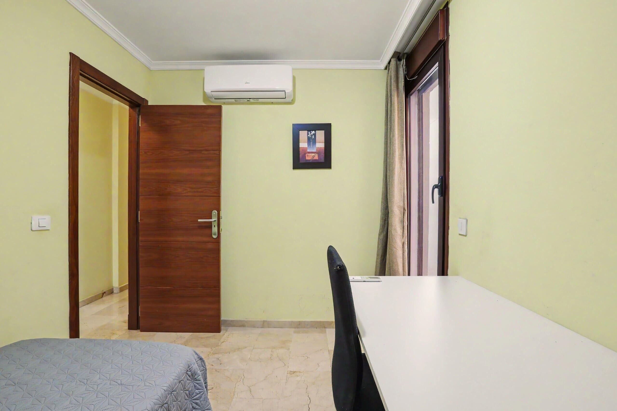 A room with a bed, a table, and a chair. There is a door, a picture on the wall, and an air conditioner on the ceiling.