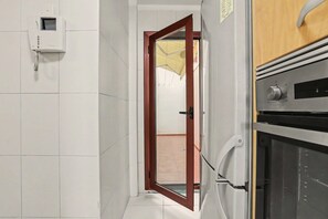 Ein schmaler Flur mit weiß gefliester Wand, einer Holztür, einem Telefon und einem Edelstahlbackofen.