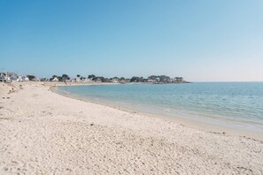 A wide sandy beach with a calm sea, a clear sky, and houses in the background.