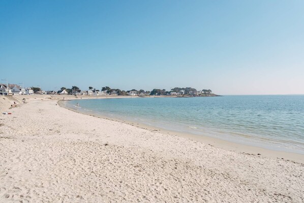 A wide sandy beach with a calm sea, a clear sky, and houses in the background.