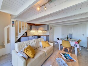A modern living room with a sofa, a dining table set for two, and a kitchen area with a staircase.
