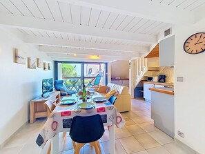 A dining area with a table set for four, a kitchen in the background, and a clock on the wall.