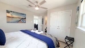 Un dormitorio con cama, ventilador de techo, pintura de una playa, armario y ventana.