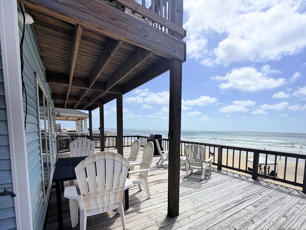 Eine Holzterrasse mit weißen Stühlen und einem Tisch, mit Blick auf einen Strand.