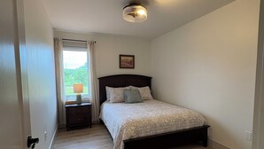 Um quarto com cama, mesa de cabeceira, luminária, luz de teto e janela com cortinas.