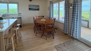Uma sala de jantar com mesa e cadeiras de madeira, uma cozinha com pia e armários, e janelas amplas com cortinas.