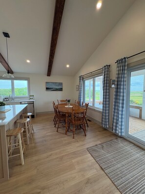 A dining area with a wooden table and chairs, a kitchen with a sink and cabinets, and large windows with curtains.