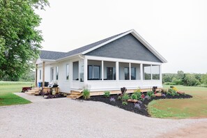 A single-story house with a covered porch, a gravel driveway, and a landscaped garden with various plants and flowers. - Brand New Cottage in Stratford-Hot Tub, incredible water view! (Stratford)