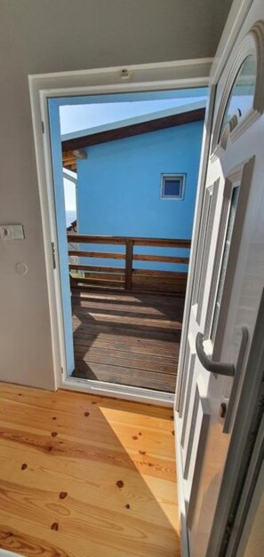 A room with a wooden floor, a door leading outside, and a view of a blue building with a balcony.
