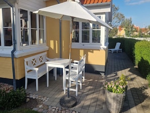 Eine Terrasse mit Tisch und Stühlen, einem weißen Sonnenschirm und einem Korbusch in Blumen.