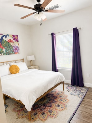 A bedroom with a bed, a ceiling fan, a window with curtains, a nightstand, a lamp, and a colorful rug. - Pass Time Retreat- 1/2 Mile to the beautiful Pass Christian Beaches- Sleeps 6 (Pass Christian)
