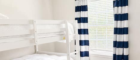 Una cama individual en literas en una habitación con ventilador de techo, suelo de madera y una ventana con cortinas a rayas.