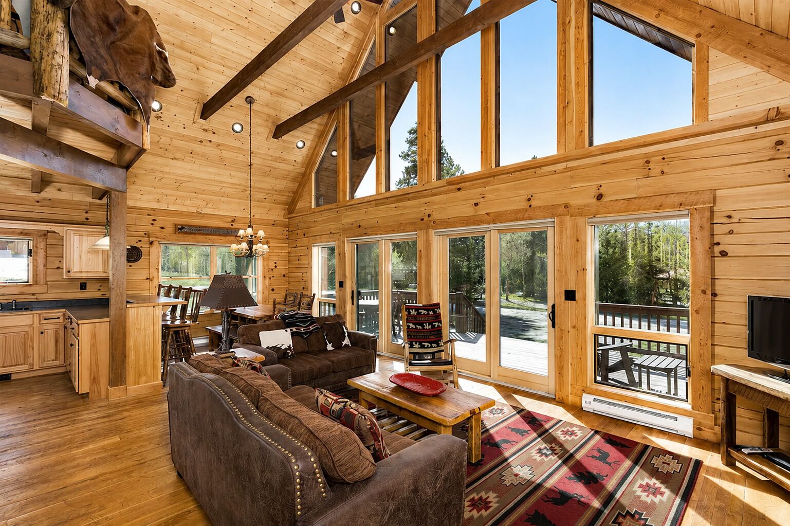 A log cabin interior with a large wooden ceiling, a cozy living area with a sofa and chairs, a dining table, and a TV.