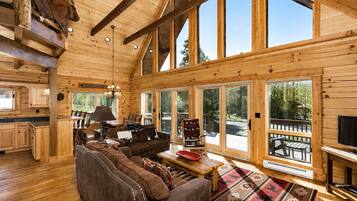 A log cabin interior with a large wooden ceiling, a cozy living area with a sofa and chairs, a dining table, and a TV.
